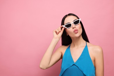 Beautiful young woman in stylish sunglasses blowing kiss on pink background. Space for text