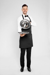 Handsome waiter holding metal tray with lid on light background