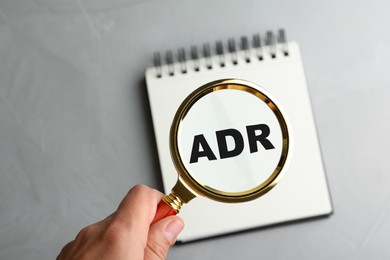 Alternative dispute resolution. Closeup of woman holding magnifier glass over abbreviation ADR in notebook at light grey table