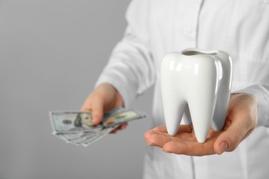 Photo of Dentist holding ceramic model of tooth and dollar banknotes on grey background, closeup. Expensive treatment