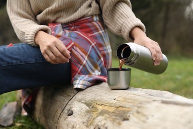 Woman pouring hot drink from metallic thermos into cup lid outdoors, closeup