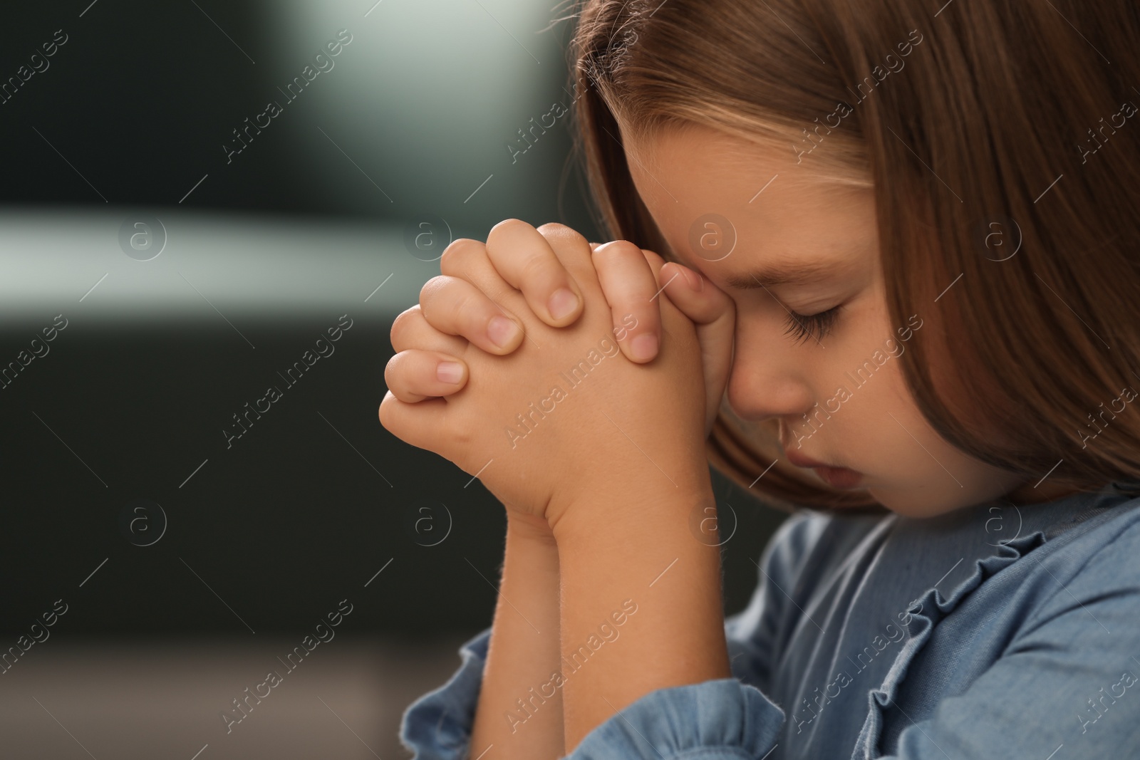 Photo of Cute little girl with hands clasped together praying on blurred background, closeup