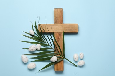 Photo of Wooden cross, painted Easter eggs and palm leaf on light blue background, flat lay