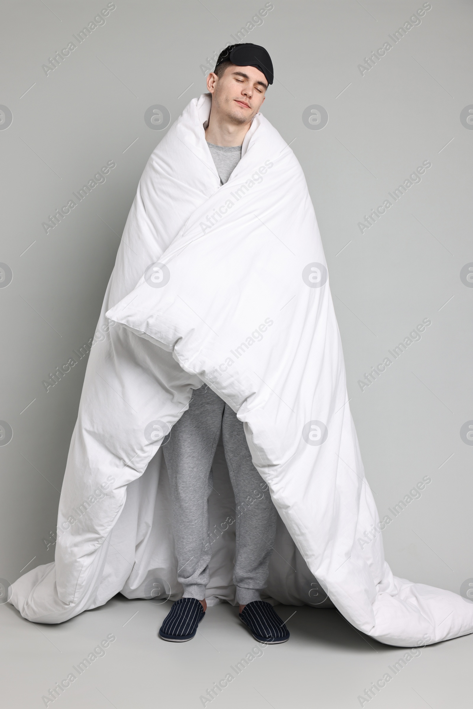 Photo of Man in pyjama and sleep mask wrapped in blanket on grey background