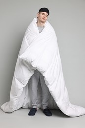 Man in pyjama and sleep mask wrapped in blanket on grey background