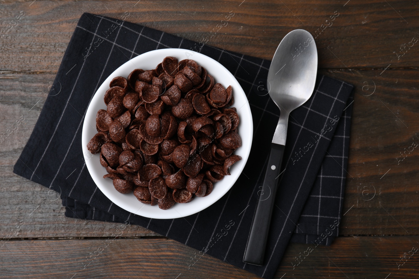 Photo of Chocolate corn flakes in bowl and spoon on wooden table, top view. Breakfast cereal