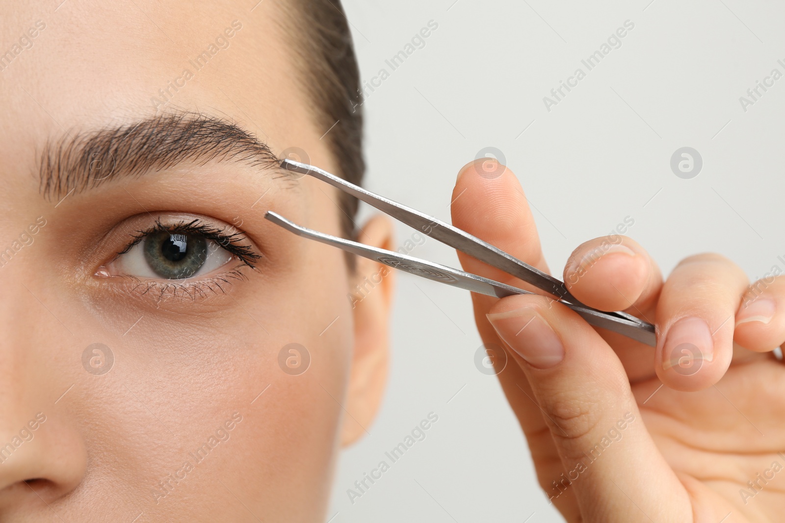 Photo of Eyebrow correction. Young woman with tweezers on light grey background, closeup