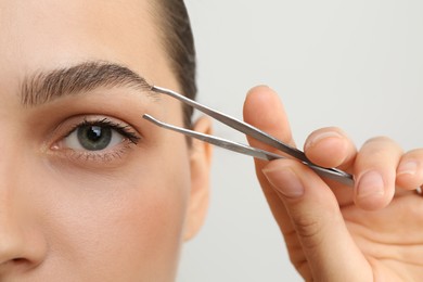 Eyebrow correction. Young woman with tweezers on light grey background, closeup