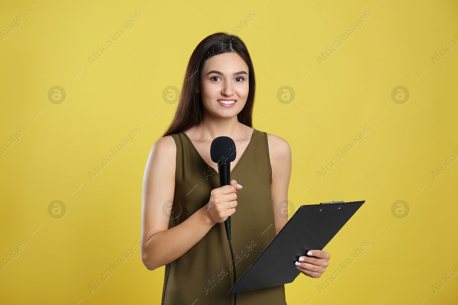 Photo of Young female journalist with microphone and clipboard on yellow background