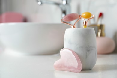 Photo of Rose quartz gua sha tool near holder with natural face rollers on white countertop in bathroom. Space for text