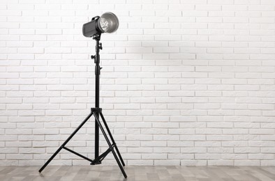 Photo of Professional studio flash light with reflector on tripod against white brick wall indoors, space for text. Photography equipment