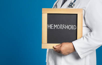 Doctor holding blackboard with word HEMORRHOID on blue background, closeup. Space for text