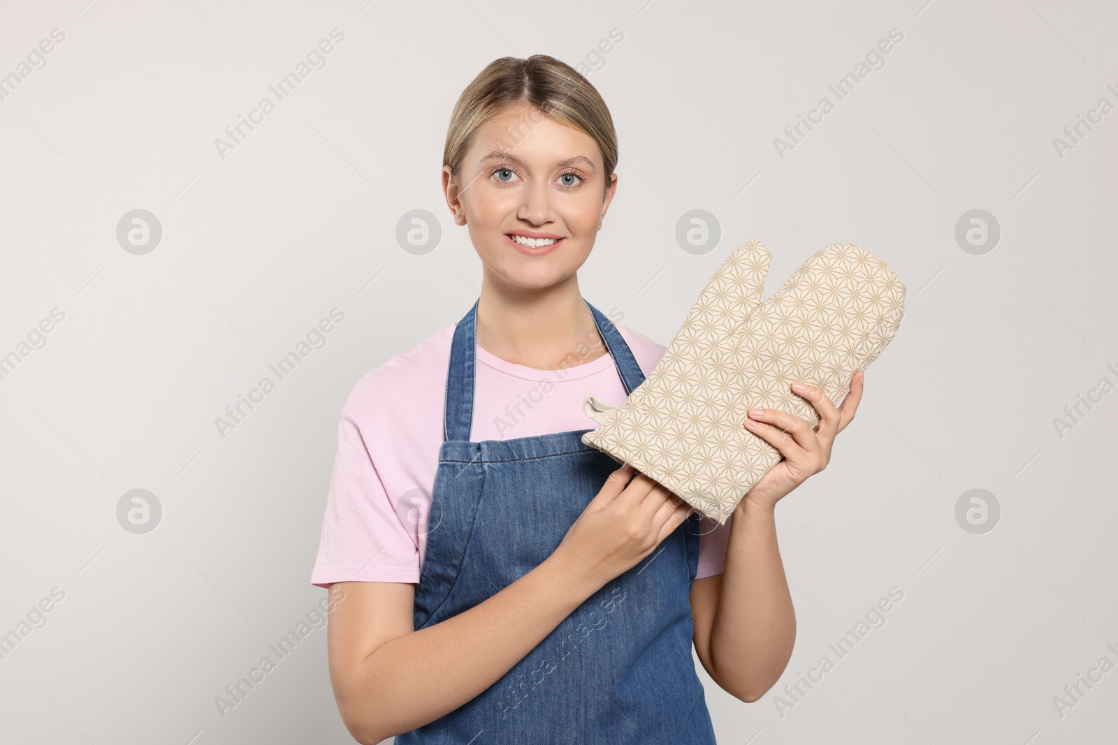 Photo of Beautiful young woman in denim apron and oven glove on light grey background