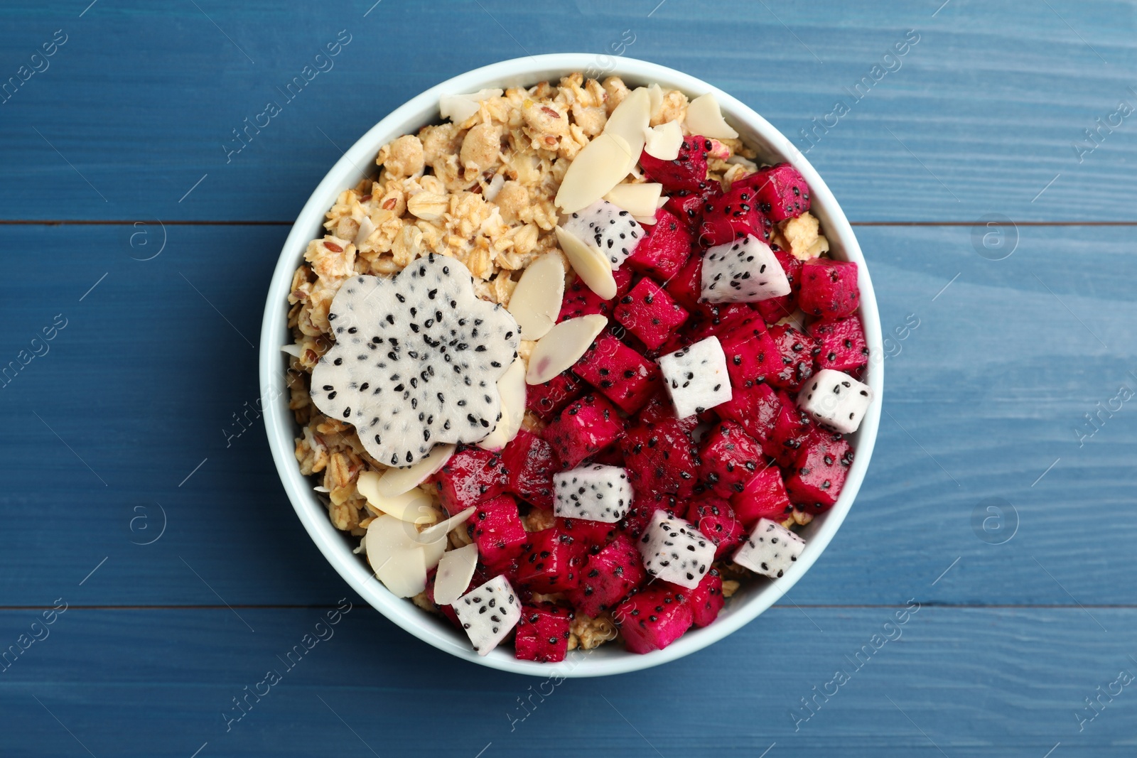 Photo of Bowl of granola with pitahaya and almond petals on blue wooden table, top view