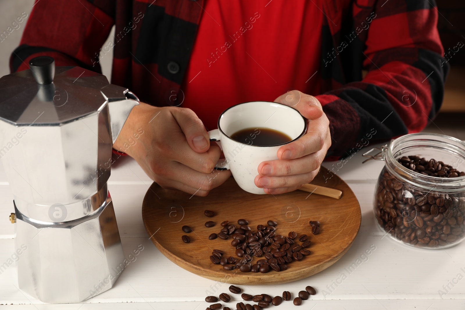 Photo of Man with cup of aromatic brewed coffee indoors, closeup. Moka pot and beans on white wooden table
