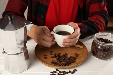 Man with cup of aromatic brewed coffee indoors, closeup. Moka pot and beans on white wooden table