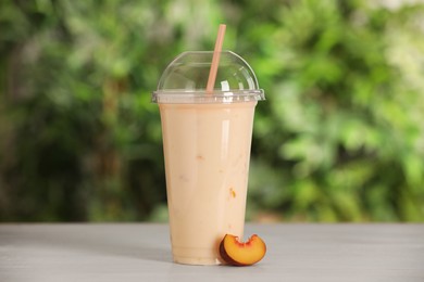 Photo of Plastic cup with tasty smoothie and slice of fresh peach on wooden table outdoors