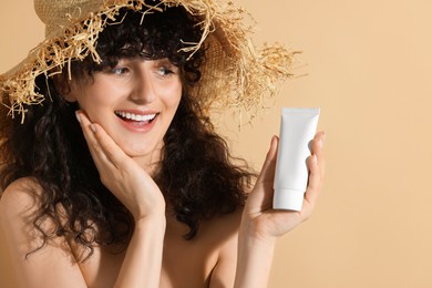 Photo of Beautiful happy woman in straw hat holding tube of sun protection cream on beige background