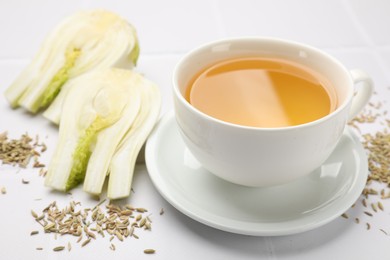 Photo of Fennel tea in cup, seeds and fresh vegetable on white tiled table, closeup