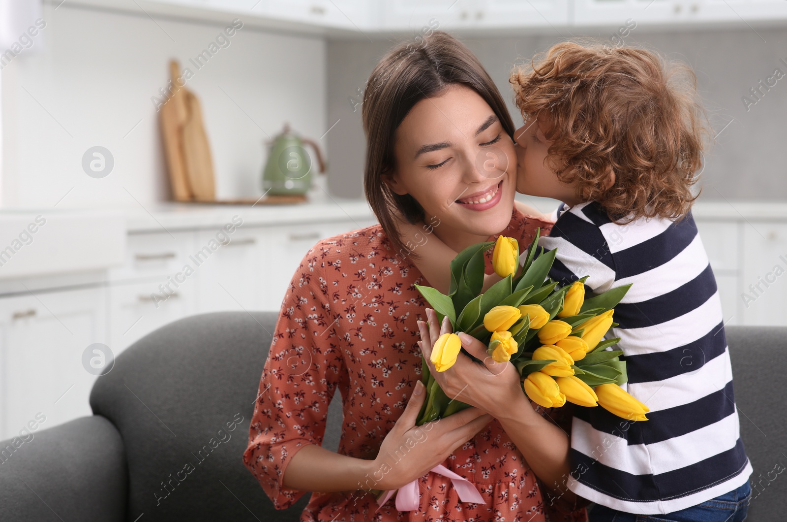 Photo of Little son kissing and congratulating his mom with Mother`s day at home. Woman holding bouquet of yellow tulips