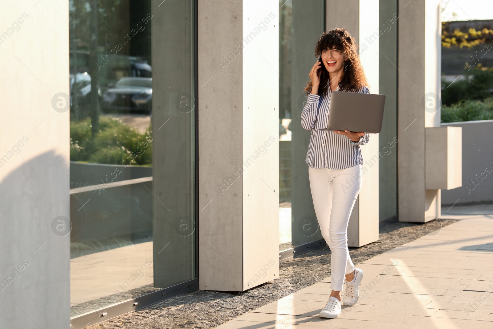 Photo of Happy young woman with modern laptop and talking on smartphone outdoors. Space for text