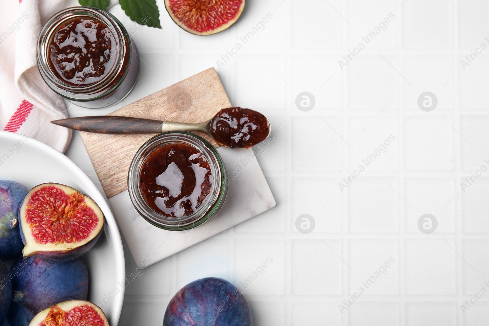 Photo of Glass jars of tasty sweet fig jam and fruits on white tiled table, flat lay. Space for text