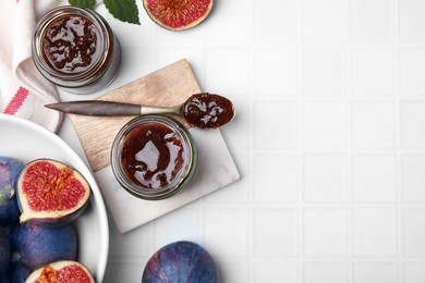 Glass jars of tasty sweet fig jam and fruits on white tiled table, flat lay. Space for text