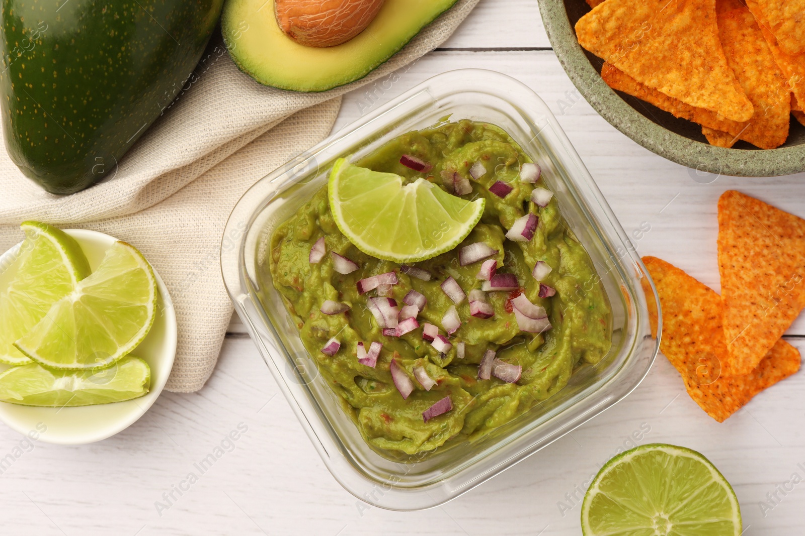 Photo of Bowl of delicious guacamole with onion, lime and nachos chips on white wooden table, flat lay