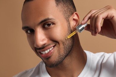 Photo of Handsome man applying cosmetic serum onto face on light brown background, closeup