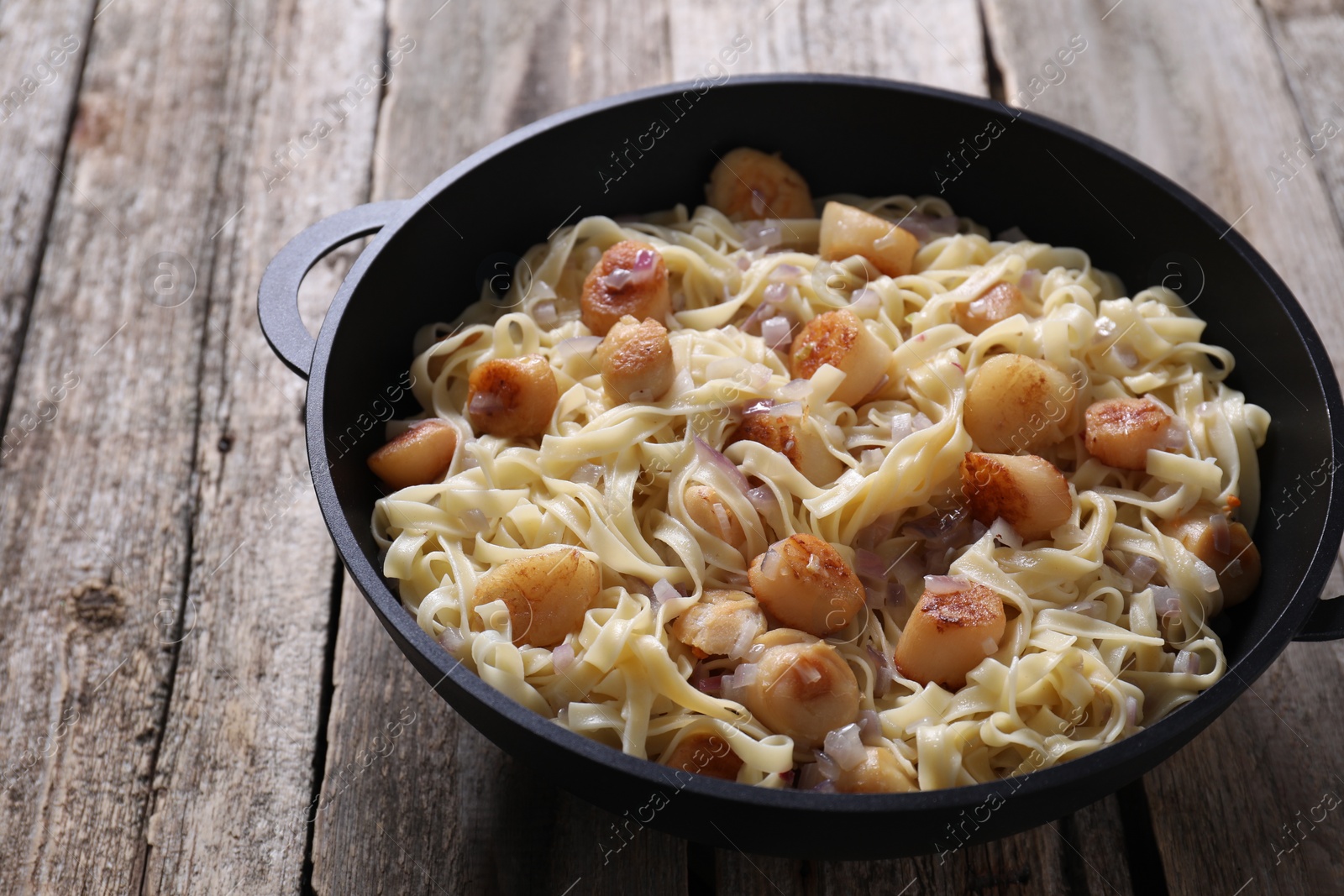 Photo of Delicious scallop pasta with onion in pan on wooden table, closeup