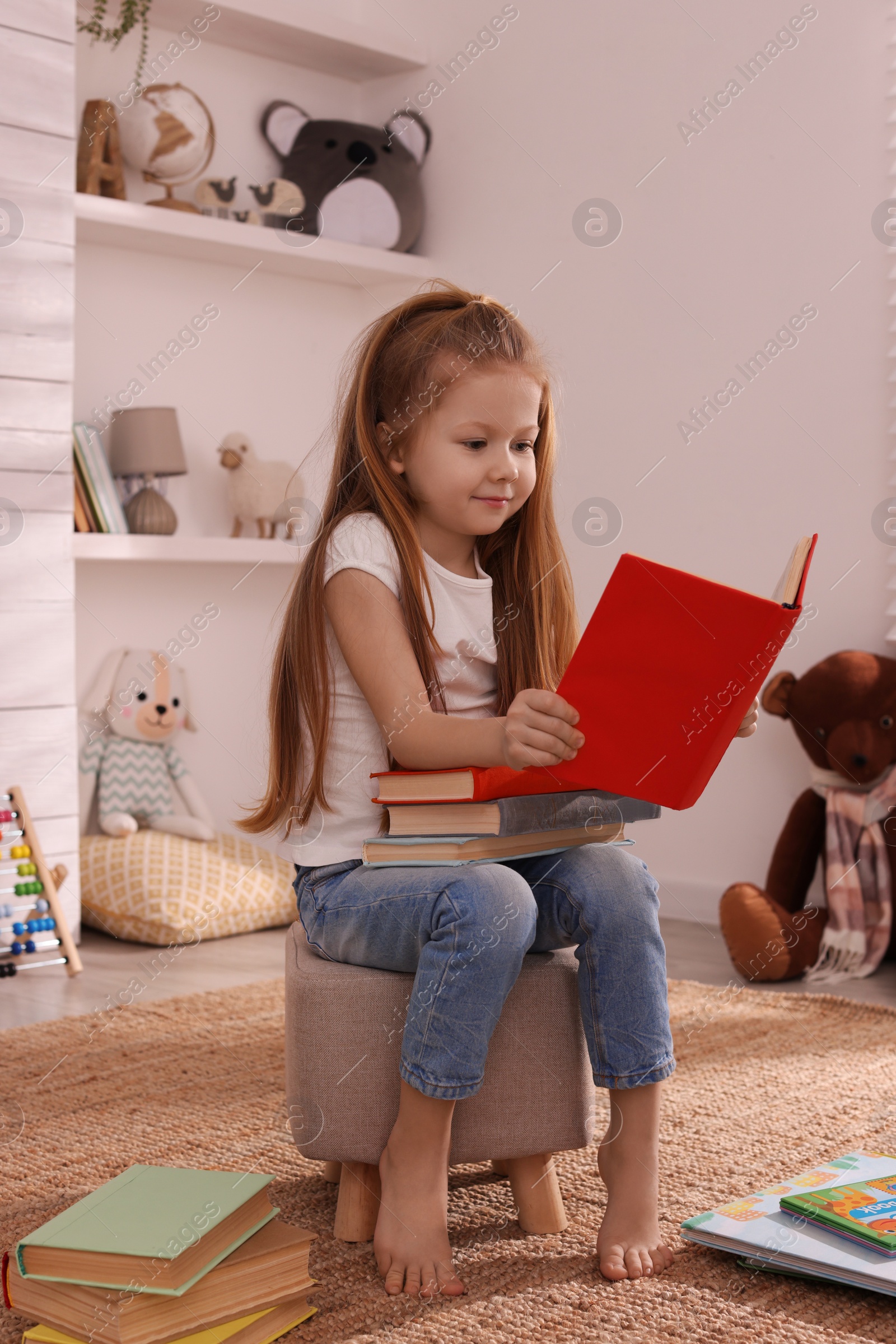 Photo of Cute little girl reading book on pouf in room