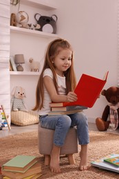 Photo of Cute little girl reading book on pouf in room