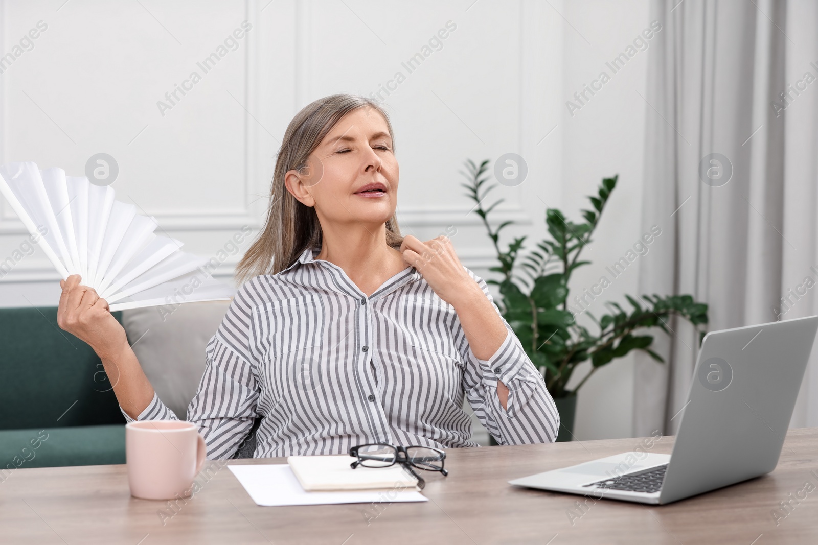 Photo of Menopause. Woman waving hand fan to cool herself during hot flash at wooden table indoors