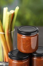 Photo of Jars of tasty rhubarb jam and stalks on table, closeup