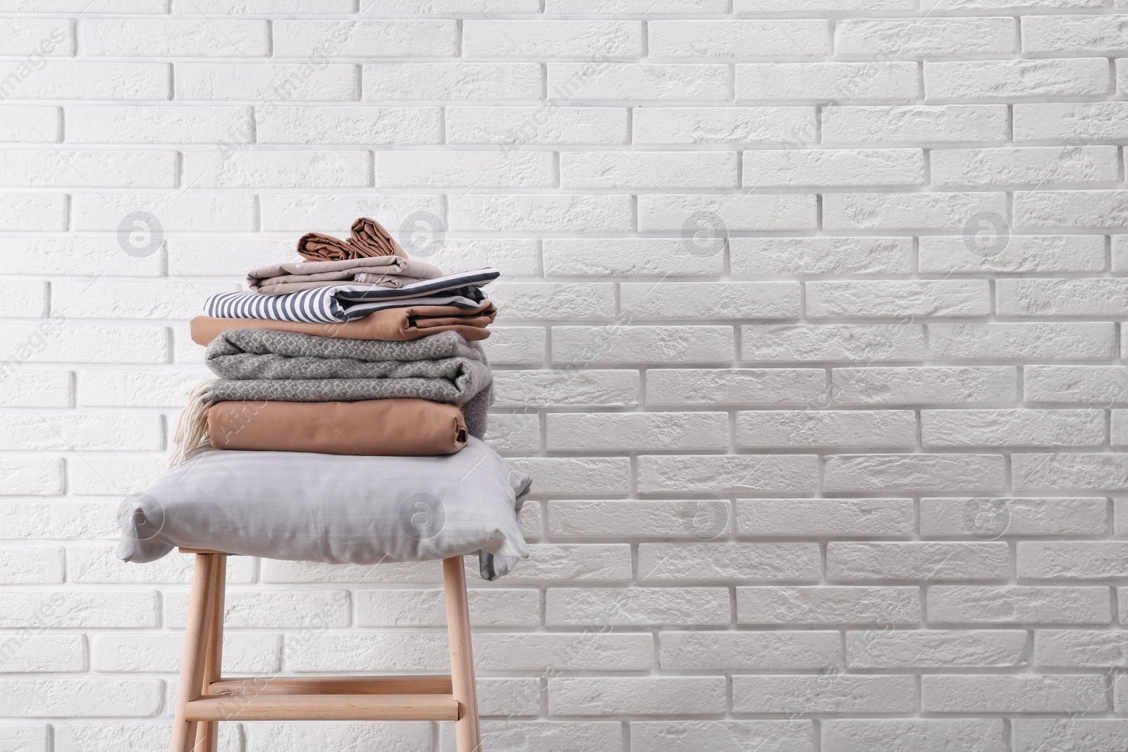 Photo of Stack of clean bed sheets and pillow on stool near white brick wall. Space for text