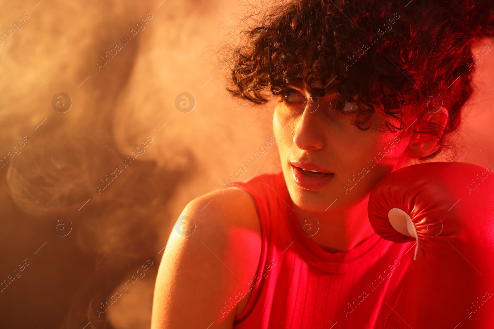 Photo of Beautiful young woman with boxing gloves on color background in neon lights and smoke, closeup. Space for text