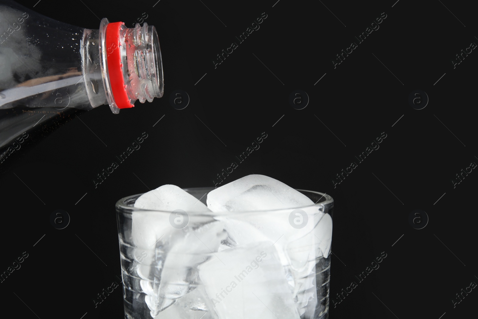 Photo of Pouring refreshing cola from bottle into glass with ice cubes on black background, closeup