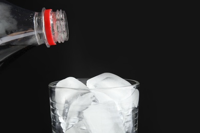 Pouring refreshing cola from bottle into glass with ice cubes on black background, closeup