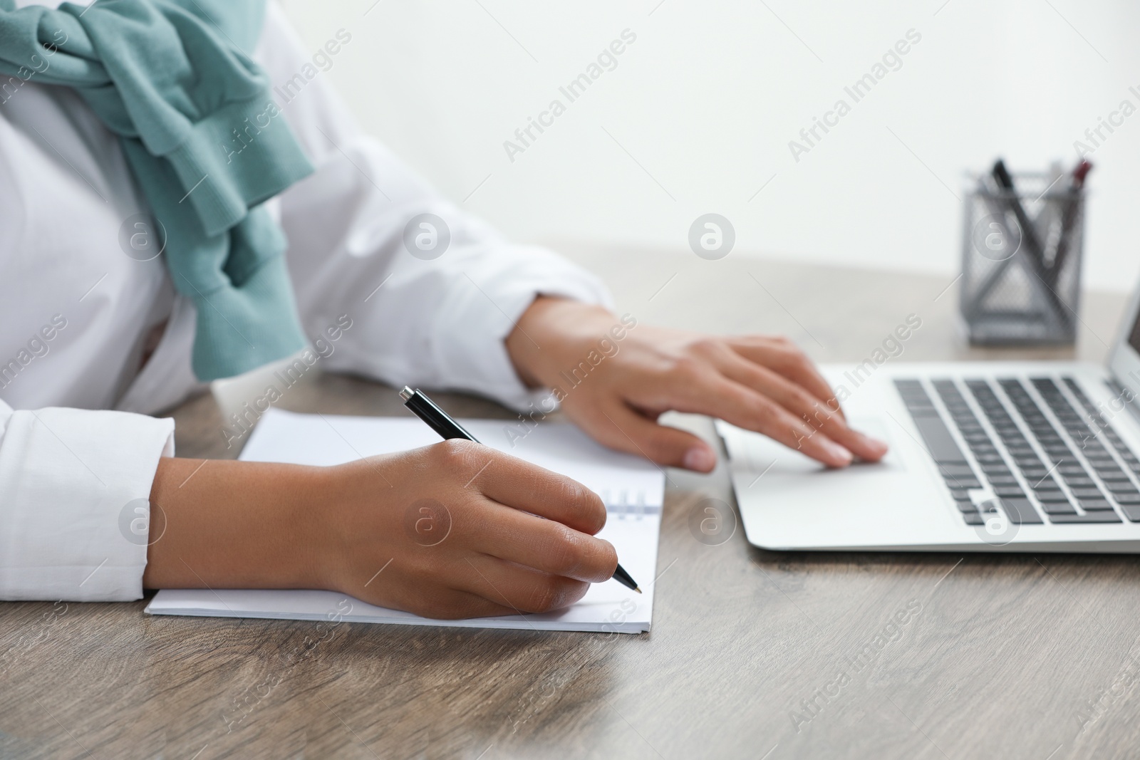 Photo of Woman writing notes while using laptop at wooden desk indoors, closeup