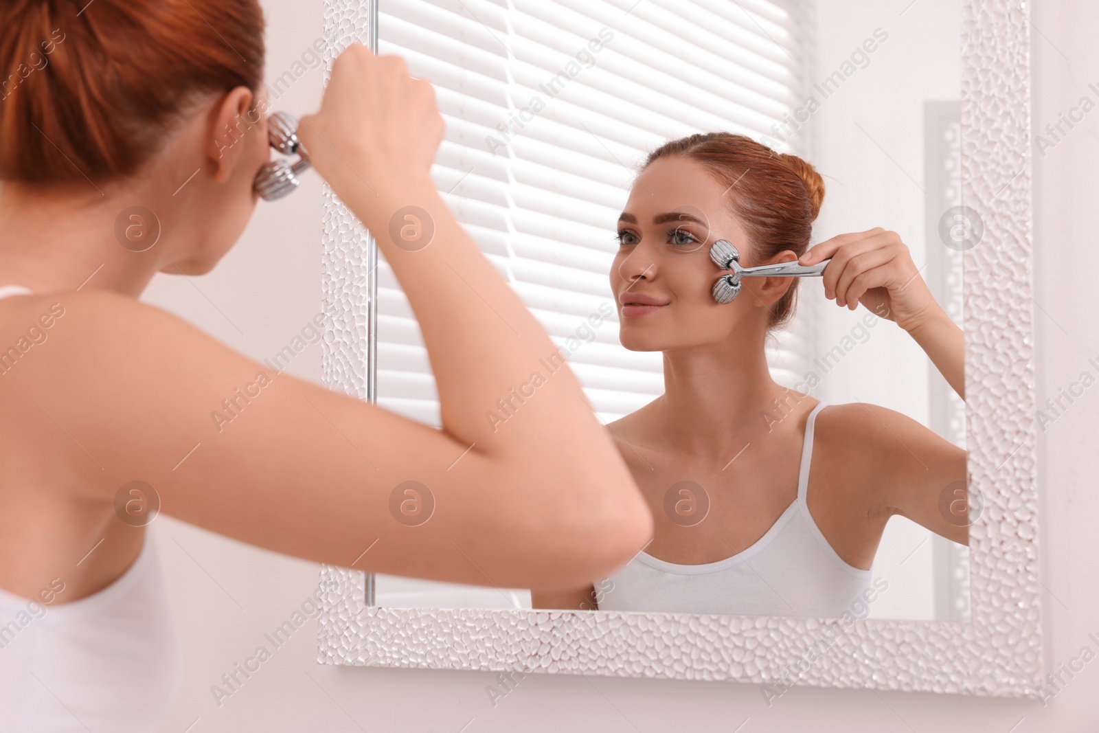 Photo of Young woman massaging her face with metal roller near mirror in bathroom