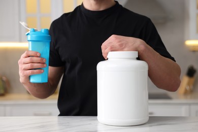 Young man with shaker of protein and powder at white marble table in kitchen, closeup