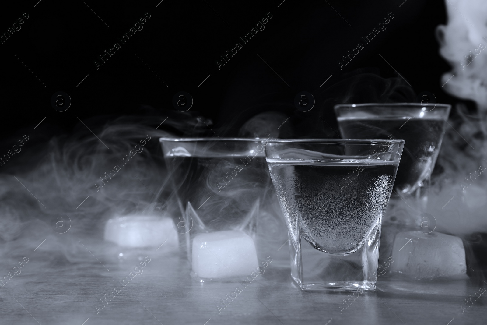 Photo of Vodka in shot glasses with ice on table against black background, closeup