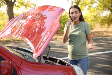 Stressed pregnant woman talking on phone near broken car outdoors