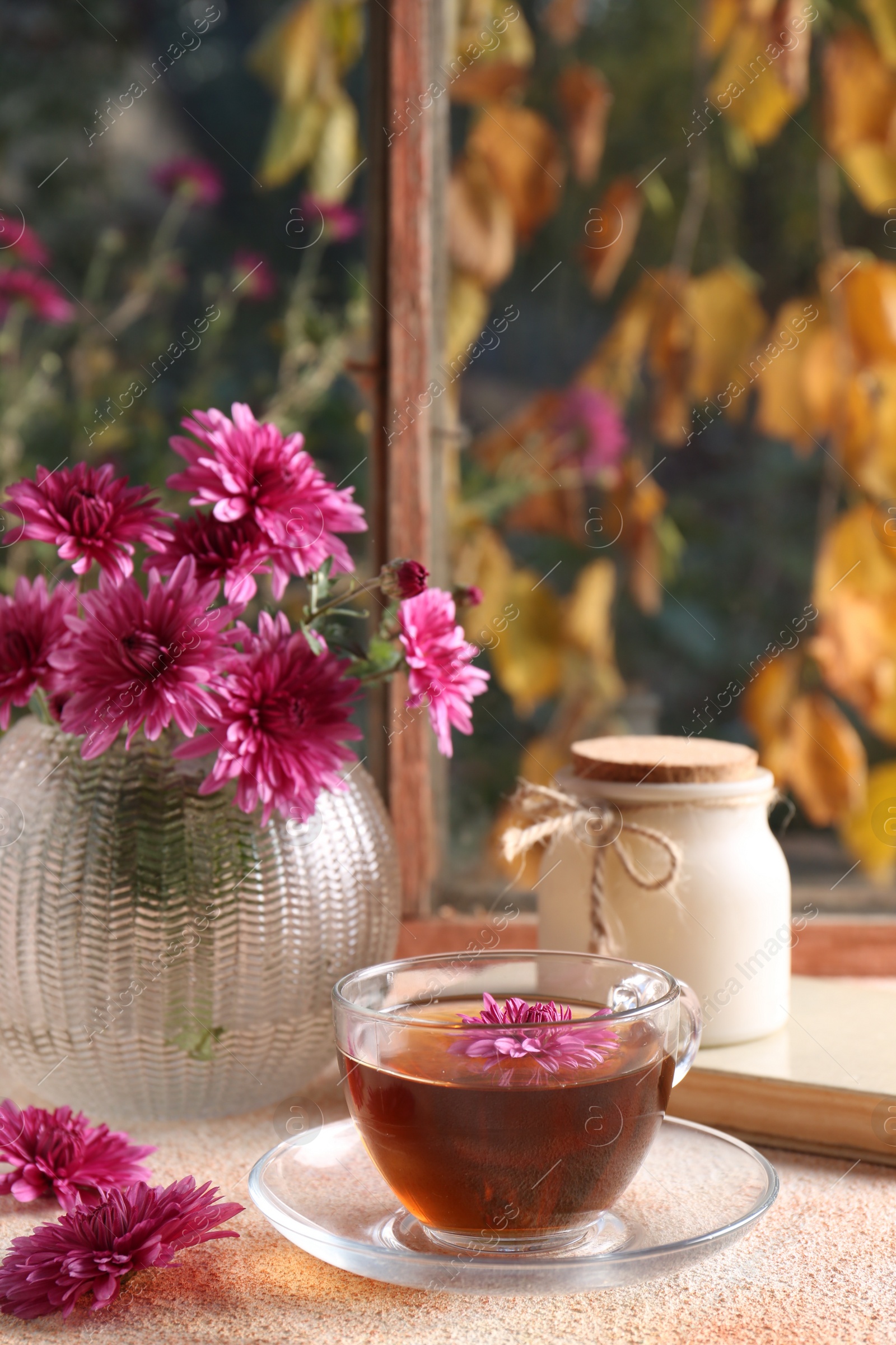 Photo of Beautiful chrysanthemum flowers in vase and cup of tea on beige textured table near window. Autumn still life