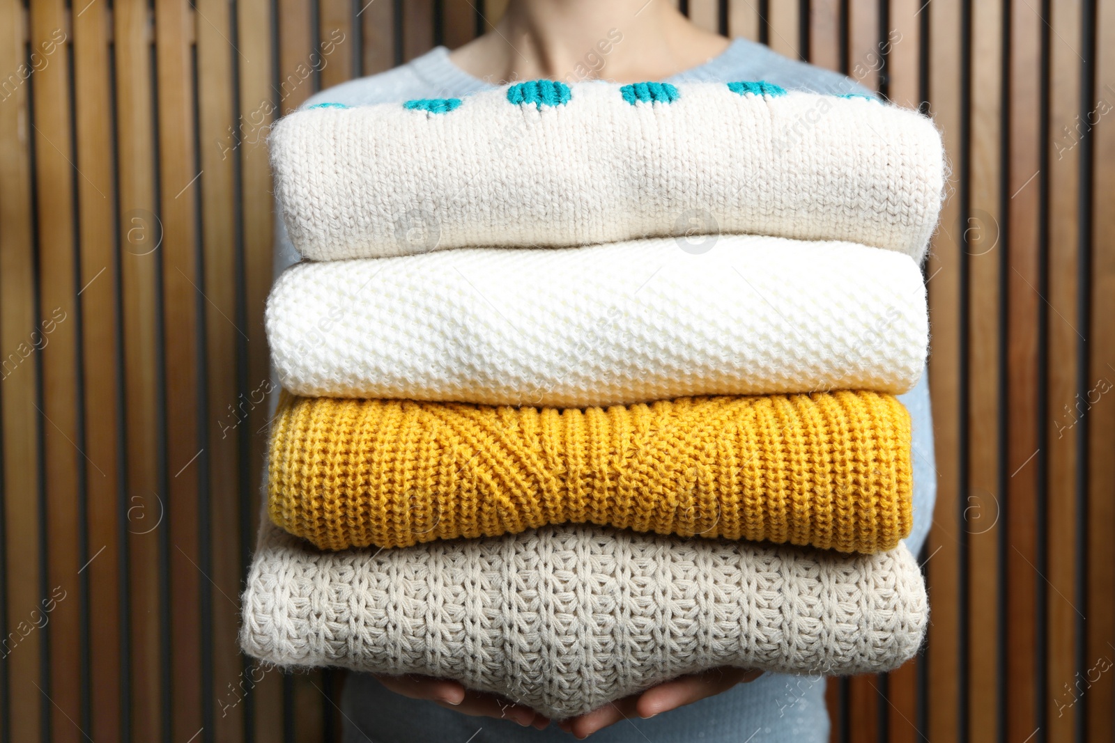 Photo of Woman holding pile of winter sweaters near wooden plank wall, closeup view