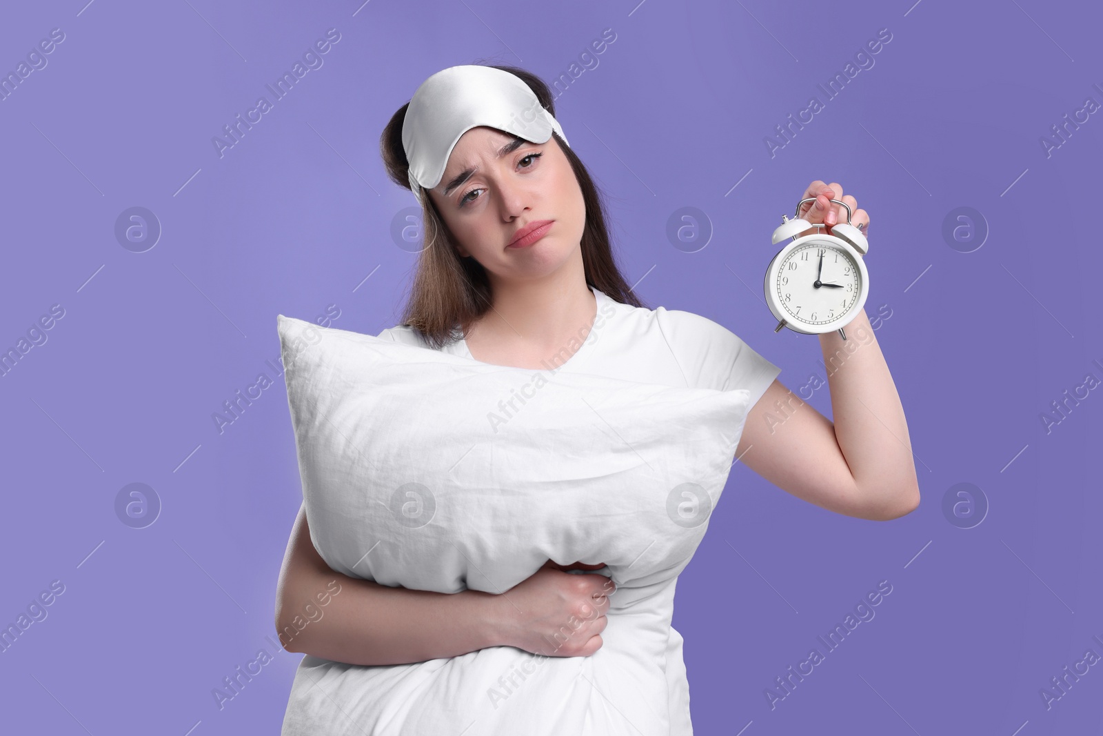 Photo of Tired young woman with sleep mask, alarm clock and pillow on purple background. Insomnia problem