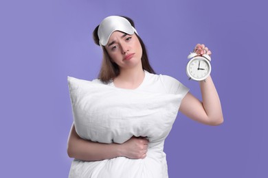 Tired young woman with sleep mask, alarm clock and pillow on purple background. Insomnia problem