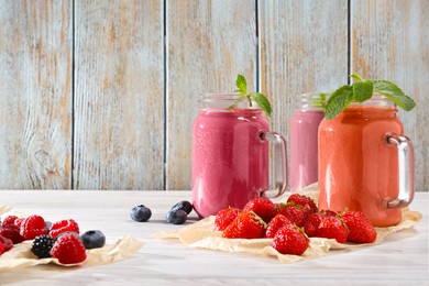 Photo of Mason jars of different berry smoothies and fresh ingredients on white wooden table. Space for text