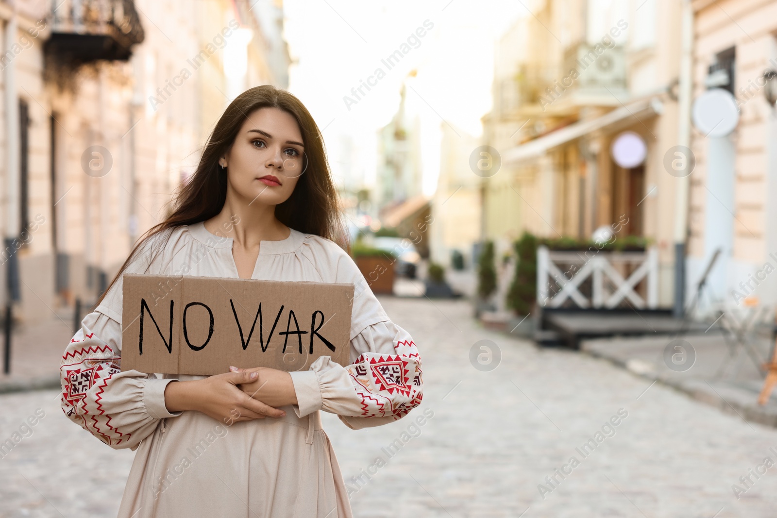 Photo of Sad woman in embroidered dress holding poster No War on city street. Space for text