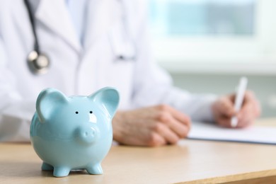 Photo of Doctor writing notes at wooden table, focus on piggy bank